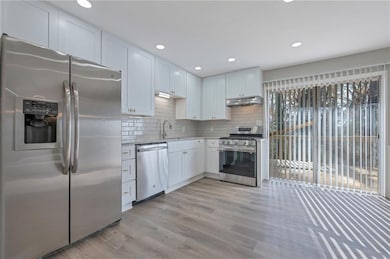 Kitchen featuring stainless steel appliances, under cabinet range hood, light wood-type flooring, decorative backsplash, and white cabinetry