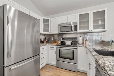 Kitchen featuring appliances with stainless steel finishes, white cabinetry, glass insert cabinets, light stone countertops, and light wood-style floors