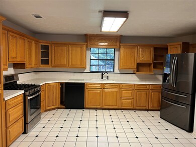 Spacious kitchen with ample wooden cabinetry, tiled flooring, and a large window. Features stainless steel appliances including a propane gas stove and double-door refrigerator.
