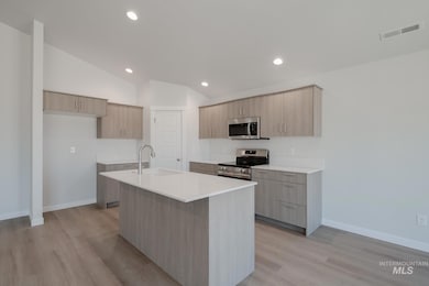 Kitchen featuring modern cabinets, appliances with stainless steel finishes, light brown cabinetry, a kitchen island with sink, and light stone counters