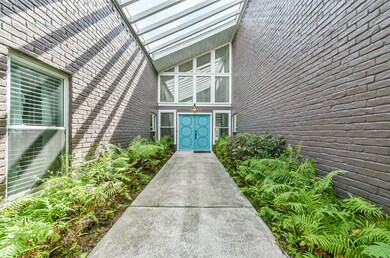 A pergola-style slatted roof accentuates the modern roofline, painted brick exterior walls, and tropical ferns that line the pathway to a double door entrance painted with a bold teal paint selection.