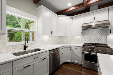 Kitchen with stainless steel appliances, under cabinet range hood, beamed ceiling, white cabinetry, and tasteful backsplash