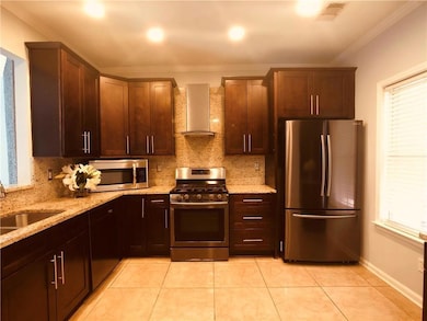 Kitchen featuring appliances with Black stainless steel finishes, wall chimney exhaust hood, ornamental molding, backsplash, and light stone counters