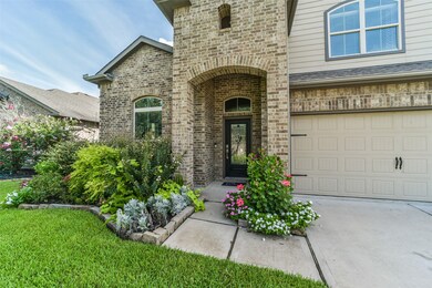 This is a well-maintained two-story brick home featuring a covered entryway and an attached two-car garage. The front yard is landscaped with lush greenery and flowering plants, adding to the home's curb appeal.