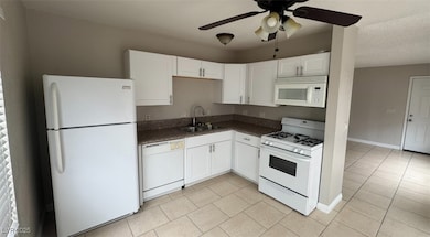 Kitchen with white appliances, dark countertops, light tile patterned flooring, ceiling fan, and white cabinetry