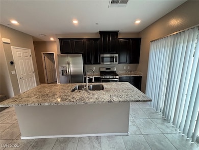 Kitchen with dark cabinets, appliances with stainless steel finishes, light stone counters, an island with sink, and recessed lighting