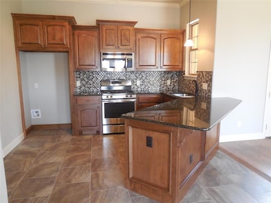 Kitchen featuring dark stone countertops, brown cabinets, appliances with stainless steel finishes, decorative light fixtures, and tasteful backsplash
