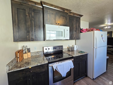 Kitchen with white appliances, dark brown cabinets, a textured ceiling, light stone countertops, and dark wood-style floors