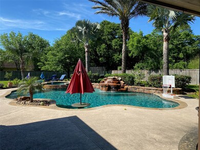 Swimming pool with hot tub and waterfall feature. Table with umbrellas holder and seating in pool. Mature palm trees. Tanning ledge by hot tub.