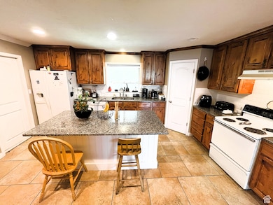 Kitchen featuring white appliances, under cabinet range hood, a kitchen breakfast bar, a sink, and dark stone counters