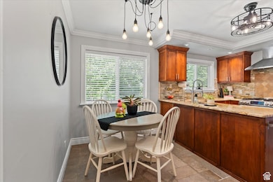 Kitchen featuring decorative backsplash, crown molding, light stone countertops, wall chimney exhaust hood, and brown cabinetry