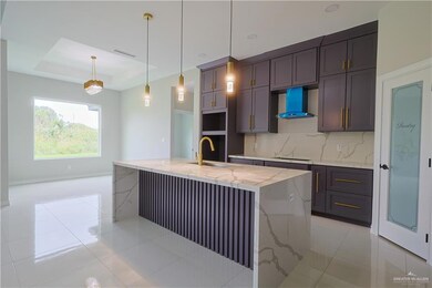 Kitchen featuring wall chimney range hood, backsplash, a center island with sink, a raised ceiling, and light stone countertops