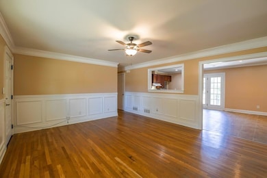 Empty room featuring wood finished floors, ornamental molding, a ceiling fan, a decorative wall, and wainscoting