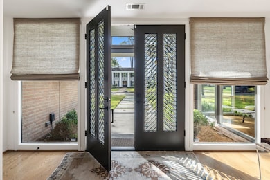A luminous foyer welcomes you inside, highlighted by the home’s original vintage double wooden doors with decorative glass detail. Floor-to-ceiling windows with custom treatments flood the space with natural light, while recessed lighting and dual walls create an ideal gallery-style entry for art or a statement console.