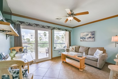 This cozy living room features a ceiling fan, comfy sofa, and stylish decor with ocean-themed art. Large glass doors lead to a balcony with a view of palm trees, providing ample natural light. The space has a relaxed, beachy vibe perfect for enjoying the outdoors.