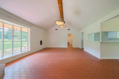 This room features a spacious, open layout with a vaulted ceiling and exposed beam. It has large windows providing natural light and views of the greenery outside. The brick flooring adds a rustic charm, and there's a pass-through to the kitchen area, enhancing the open feel.