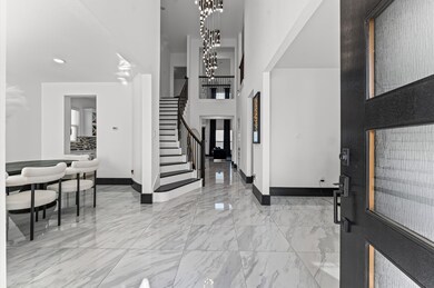 Elegant entryway features high ceilings, a modern chandelier, and sleek marble flooring. The open layout leads to a graceful staircase and a dining area with stylish seating. Natural light enhances the contemporary design.