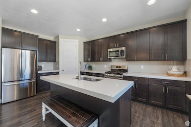 Kitchen featuring dark brown cabinets, stainless steel appliances, backsplash, a kitchen island with sink, and dark wood-style flooring