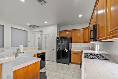 Kitchen featuring tile counters, brown cabinets, black appliances, recessed lighting, and a kitchen island with sink