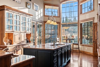 Kitchen with dark cabinetry, decorative light fixtures, an island with sink, light stone countertops, and a towering ceiling