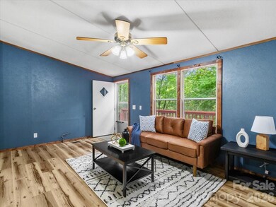 Living room with hardwood flooring, blue walls, picture windows overlooking trees, and ceiling fan. - 2271 Gouges Creek Rd, Spruce Pine, NC 28777