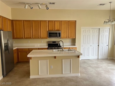 Kitchen featuring appliances with stainless steel finishes, brown cabinets, tile countertops, hanging light fixtures, and an island with sink