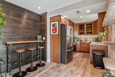 Kitchen with light stone countertops, brown cabinetry, stainless steel appliances, glass insert cabinets, and light wood-type flooring