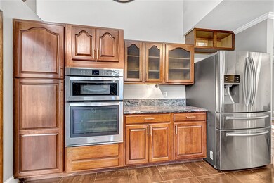 Kitchen featuring dark stone counters, appliances with stainless steel finishes, brown cabinetry, crown molding, and glass insert cabinets