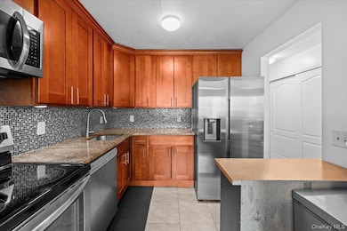 Kitchen featuring appliances with stainless steel finishes, brown cabinetry, decorative backsplash, light tile patterned floors, and light stone counters