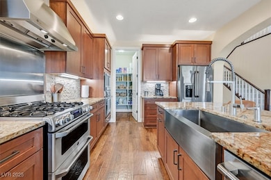Kitchen featuring appliances with stainless steel finishes, wall chimney range hood, light stone countertops, brown cabinets, and light wood-style flooring