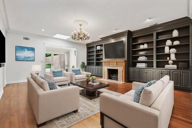 Spacious 23x17 Den has Gas Fireplace w/Carved Mantel, Wall-to-Wall Built-In Bookcases & Cabinets, HW Floors, Elegant Crown Molding, Chandelier, & Access to Sunroom Through Wide Cased Opening — Perfect for Entertaining! This photo is virtually staged.