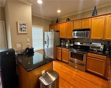 Kitchen with stainless steel appliances, light wood-style floors, brown cabinetry, backsplash, and dark stone countertops