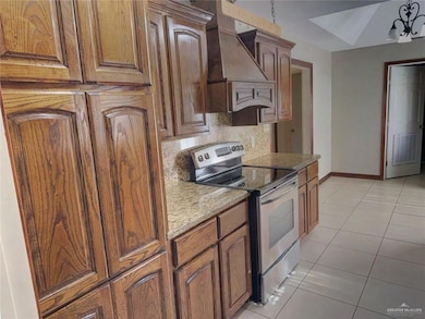 Kitchen with stainless steel electric range, backsplash, custom exhaust hood, light tile patterned flooring, and brown cabinetry