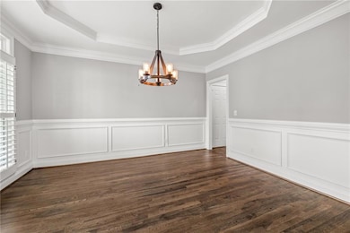 Empty room with a raised ceiling, a chandelier, healthy amount of natural light, dark wood-style flooring, and wainscoting