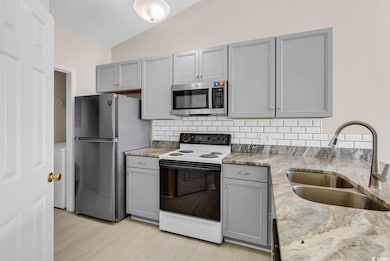 Kitchen with gray cabinets, appliances with stainless steel finishes, backsplash, light stone counters, and vaulted ceiling
