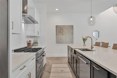 Kitchen with white cabinetry, appliances with stainless steel finishes, light stone counters, wall chimney range hood, and pendant lighting