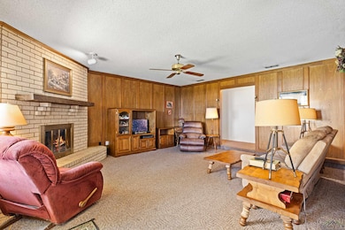 Living room with wooden walls, a ceiling fan, carpet, a textured ceiling, and a brick fireplace