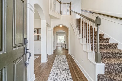 A covered front porch with a 6-panel solid wood door opens to reveal a grand foyer with a soaring two-story ceiling, a staircase with sculpted carpeting, wood-look tile floors, gently arched doorways, and neutral paint.