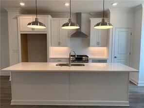 Kitchen featuring wall chimney exhaust hood, crown molding, an island with sink, light countertops, and white cabinetry
