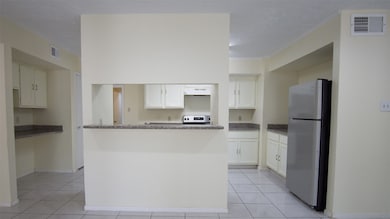 The view from the living room highlights the bright kitchen, featuring a breakfast bar, white cabinets, granite countertops, and a stainless steel refrigerator. The kitchen's tile flooring and open layout create a spacious feel.