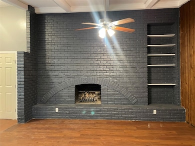 Unfurnished living room with beam ceiling, wood finished floors, a fireplace, a ceiling fan, and brick wall