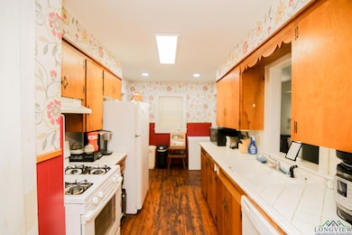 Kitchen with white appliances, wallpapered walls, a sink, tile countertops, and dark wood-type flooring