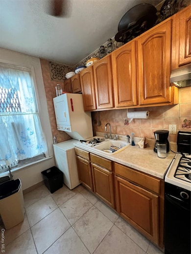 Kitchen with brown cabinetry, backsplash, light countertops, stacked washer and clothes dryer, and stove