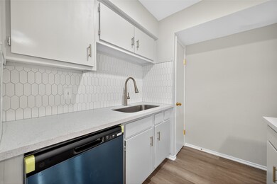 This kitchen features modern white cabinetry, a sleek stainless steel sink, and a stylish hexagonal tile backsplash. The countertop is complemented by a stainless steel dishwasher, and the space has a clean, minimalist look with a warm wood floor.