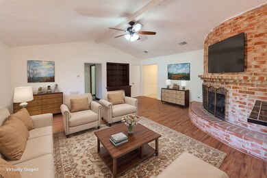 This spacious living room features a cozy, curved brick fireplace and vaulted ceilings with a ceiling fan. The room is elegantly staged with neutral-toned furniture and a large area rug, creating a warm and inviting atmosphere. Natural light enhances the open feel, and built-in shelving adds functionality.