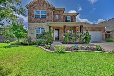 This inviting two-story brick home features a well-maintained front yard with lush landscaping and vibrant flowers. It has a covered porch, a two-car garage, and charming stone accents, all set against a clear blue sky.