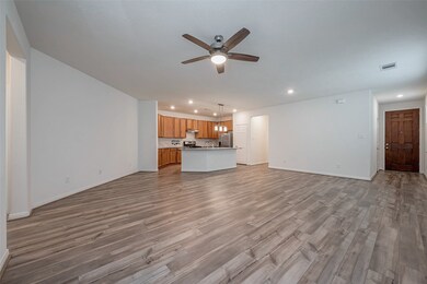 This spacious open-plan living area features wood-style flooring, a ceiling fan, and ample natural light. The modern kitchen has wooden cabinetry, stainless steel appliances, and a large island with pendant lighting, perfect for entertaining.