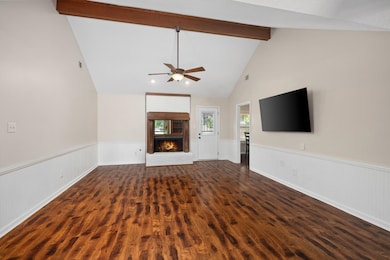 Unfurnished living room with beamed ceiling, wood finished floors, a ceiling fan, wainscoting, and a brick fireplace
