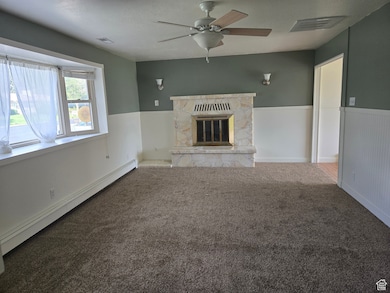 Unfurnished living room with a baseboard radiator, carpet floors, a fireplace, a wainscoted wall, and a ceiling fan