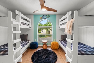 This cozy bedroom features two sets of white bunk beds, a large arched window for natural light, and a ceiling fan. The room has wood flooring and is accented with a soft blue wall, creating a welcoming space for children.